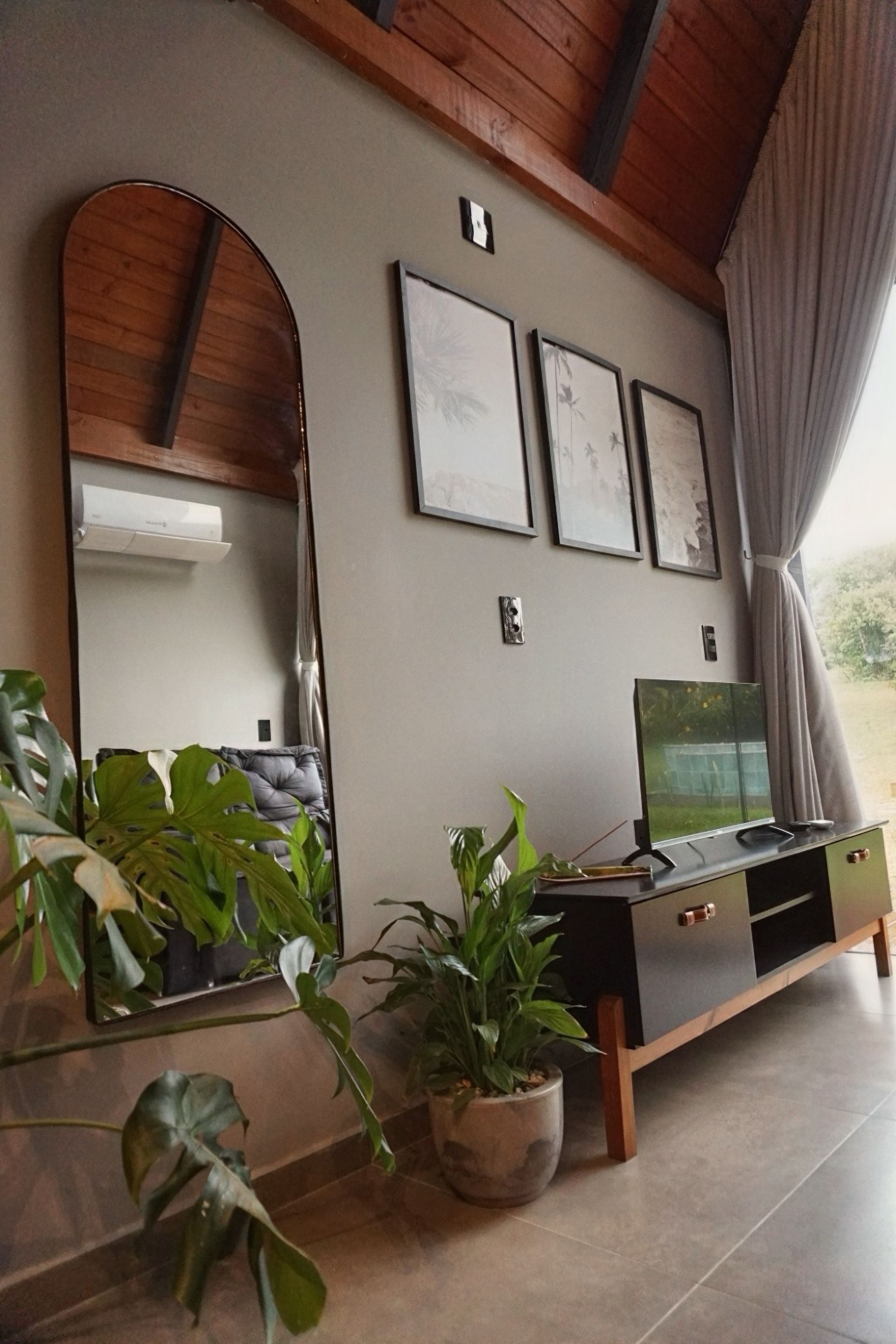 Sala de estar aconchegante com espelho grande, plantas em vasos, obras de arte na parede e uma TV em um console de madeira.