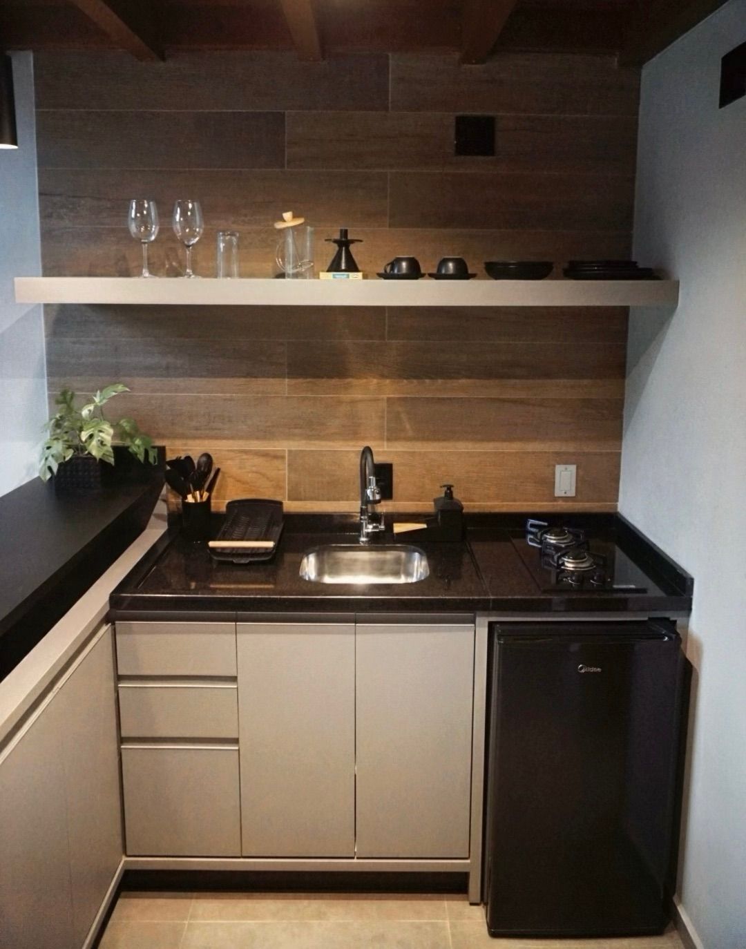 Cozinha compacta e moderna com painel de madeira na parede, pia, bancada preta, prateleira aberta e frigobar.
