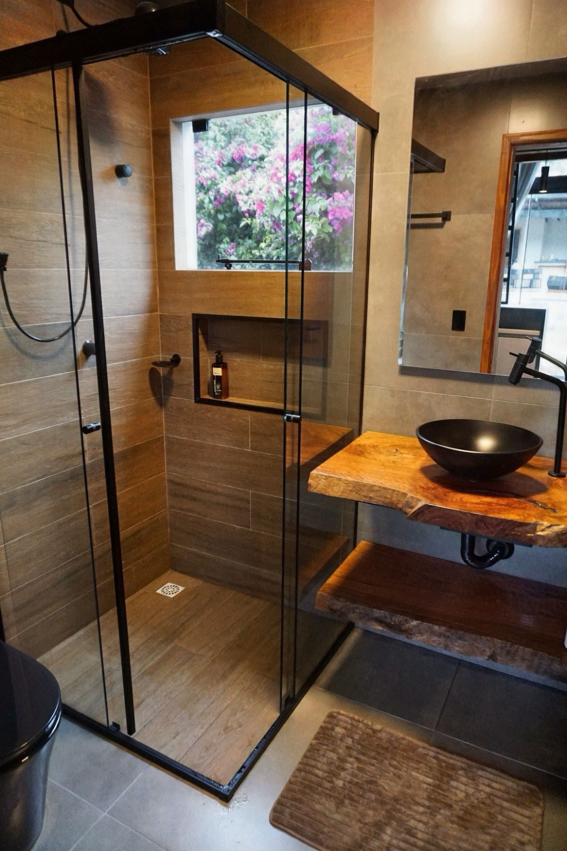 Banheiro moderno com box de vidro, bancada de madeira, pia preta e janela com flores cor-de-rosa.