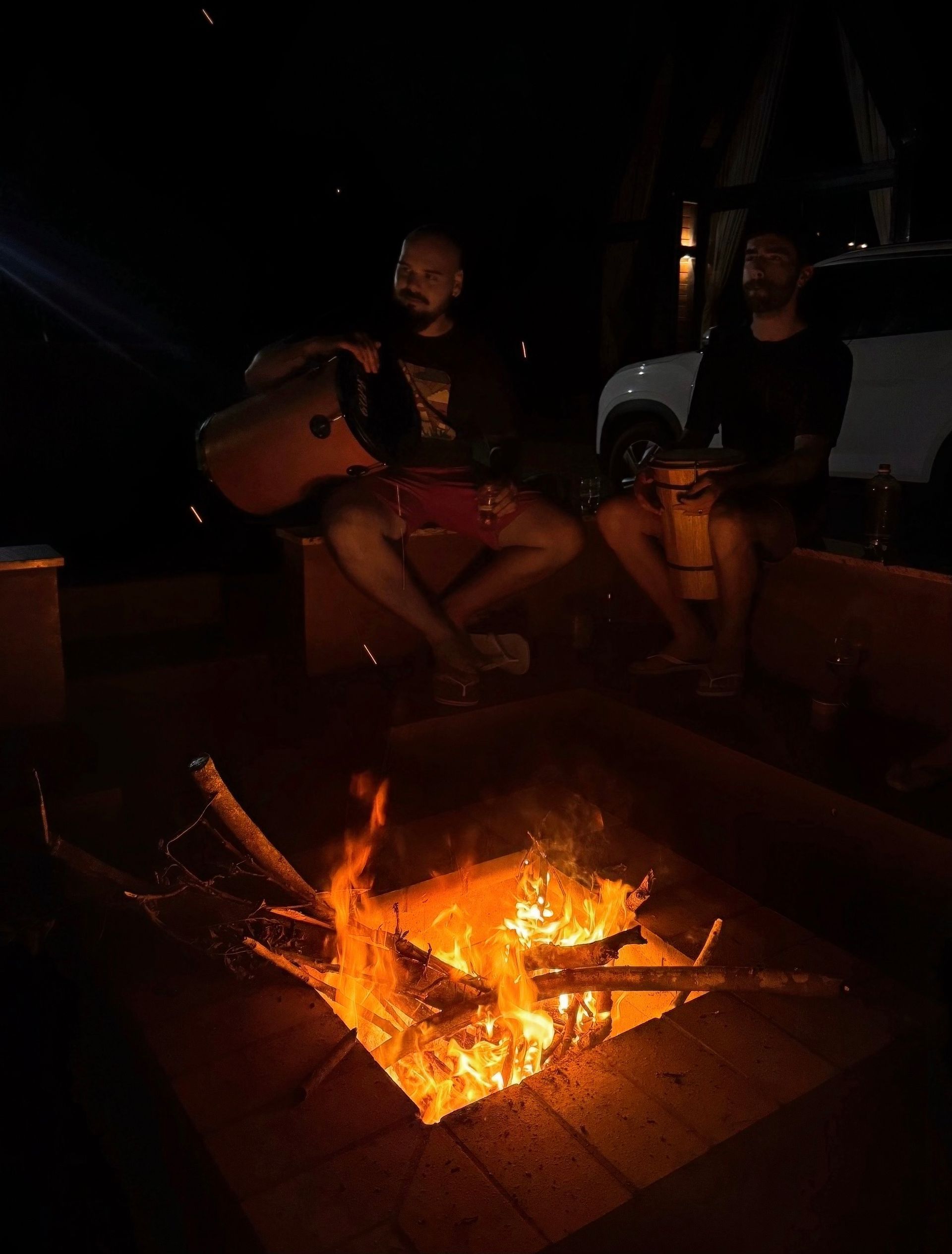Duas pessoas sentam-se ao redor de uma fogueira brilhante à noite, uma delas segurando um violão.
