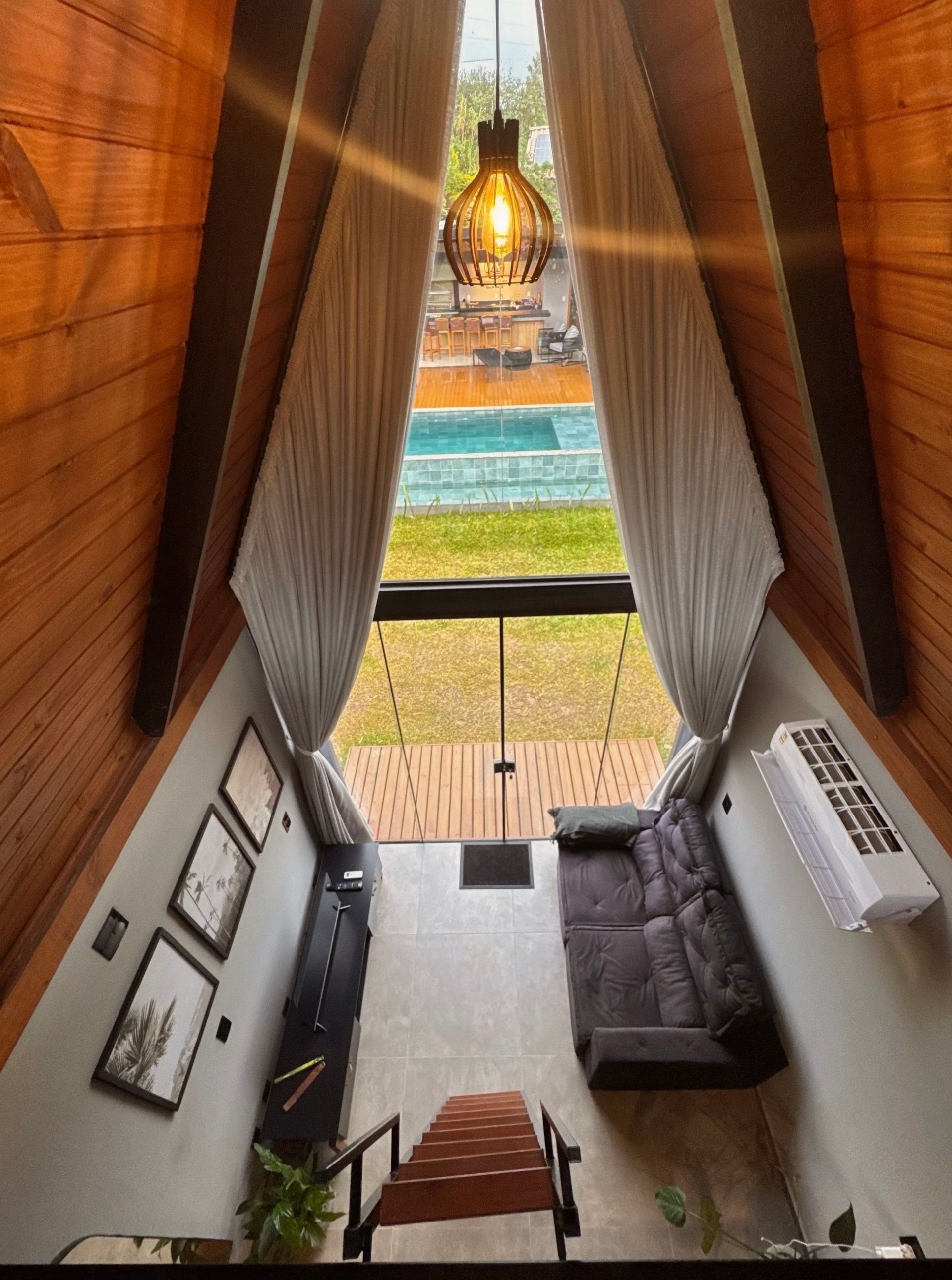 Uma sala de estar estreita com tetos altos de madeira, uma luminária pendente, sofá, escadas e uma piscina visível através de cortinas.