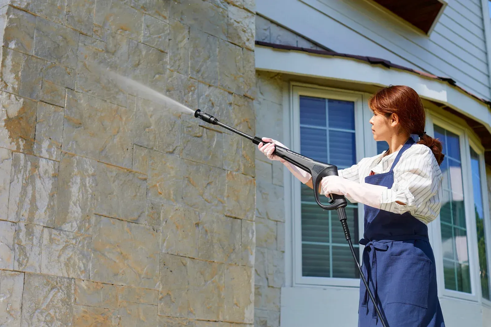 Woman in apron power washing a stone wall next to a house with windows.