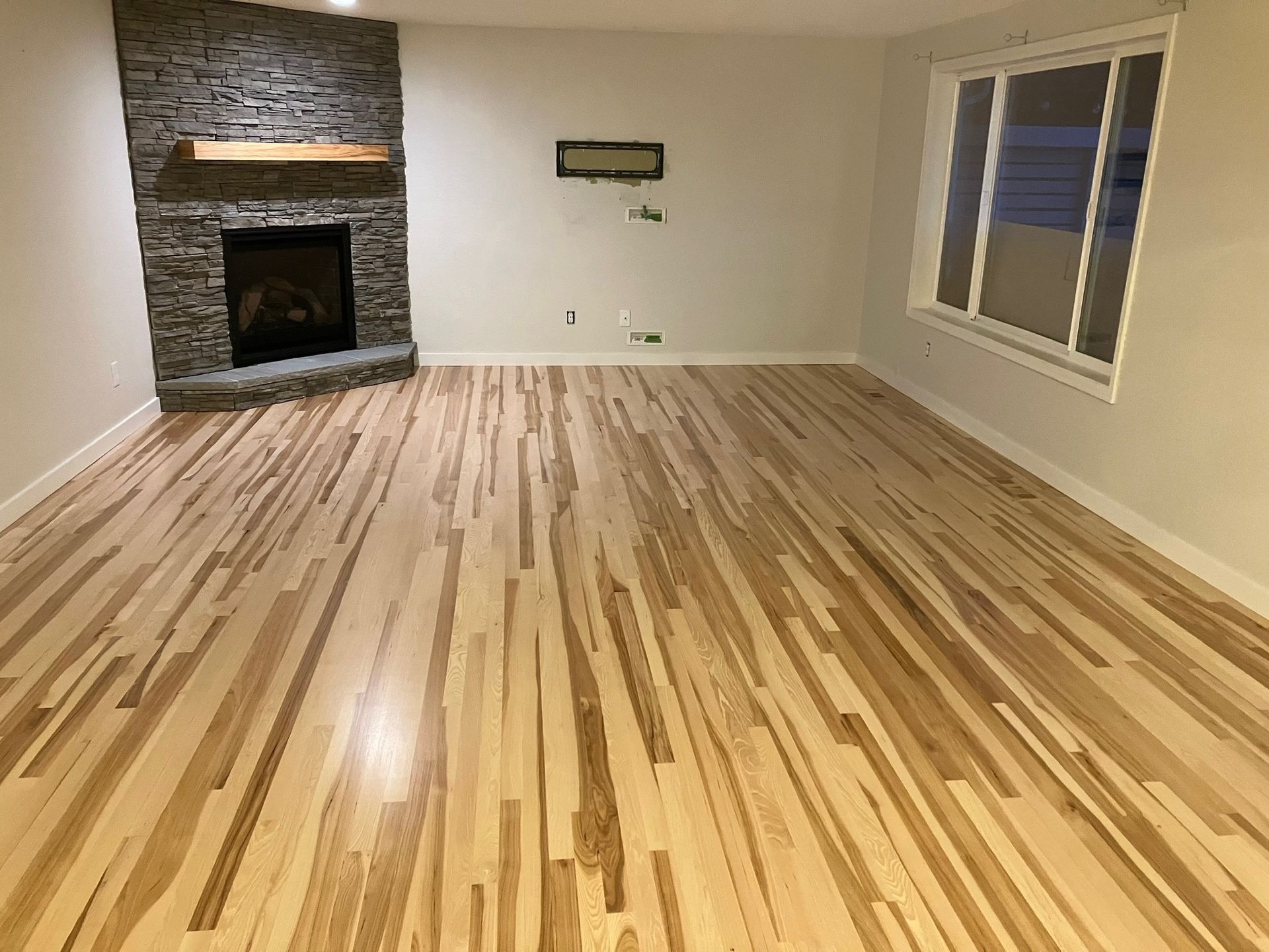 A modern, empty living room with light-colored hardwood floors, white walls, a stone corner fireplace, and a large window.