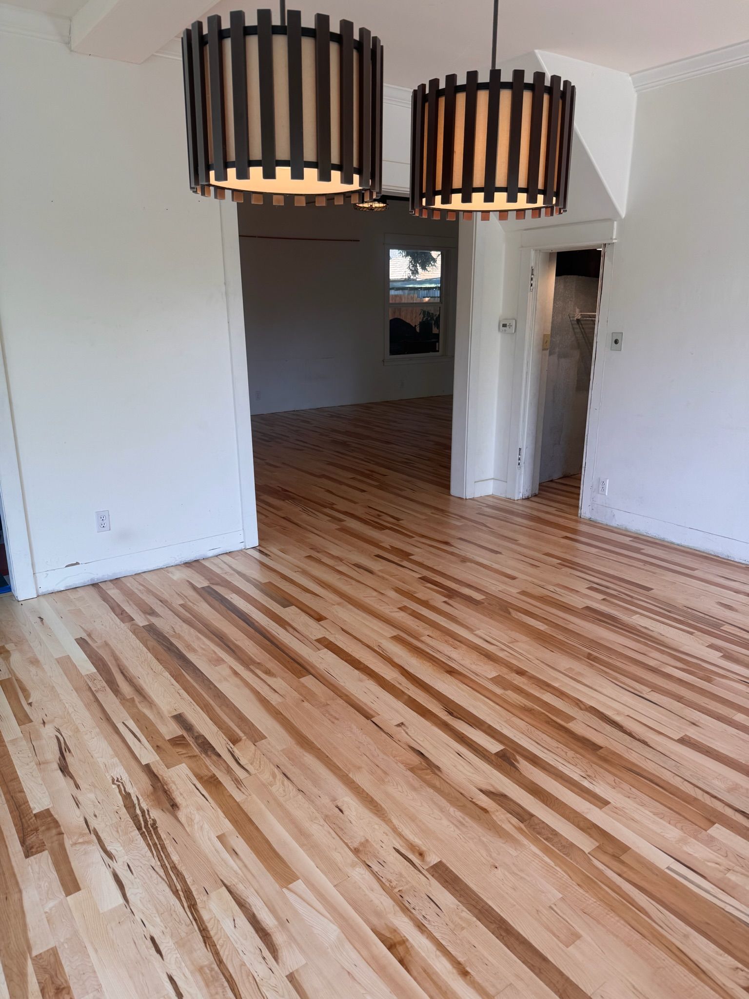 Light-colored hardwood flooring flows between two rooms under two brown, cylindrical, slatted pendant light fixtures.