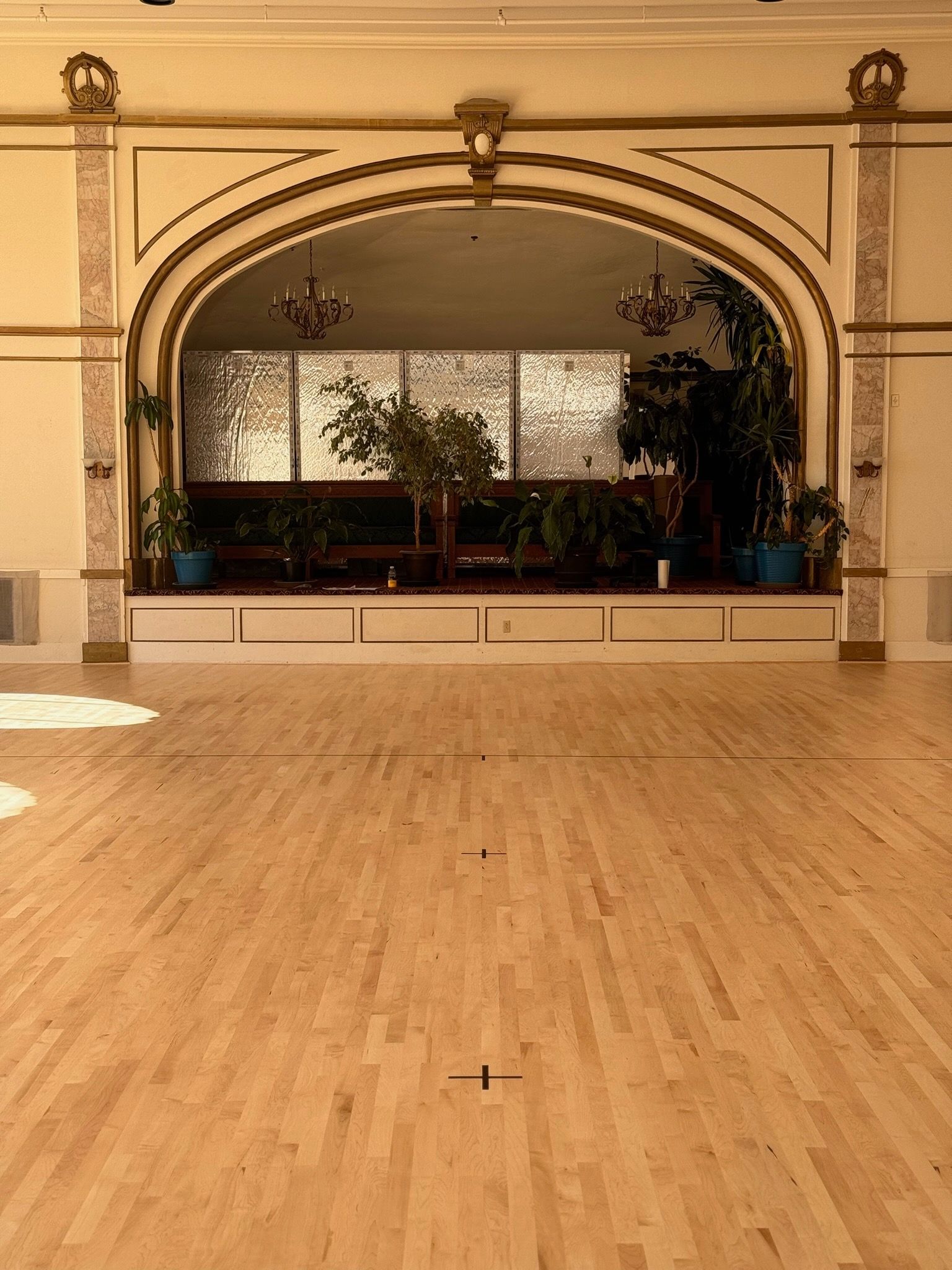 A spacious ballroom with a large wooden floor and a wide, arched stage decorated with plants and gold-trimmed columns.