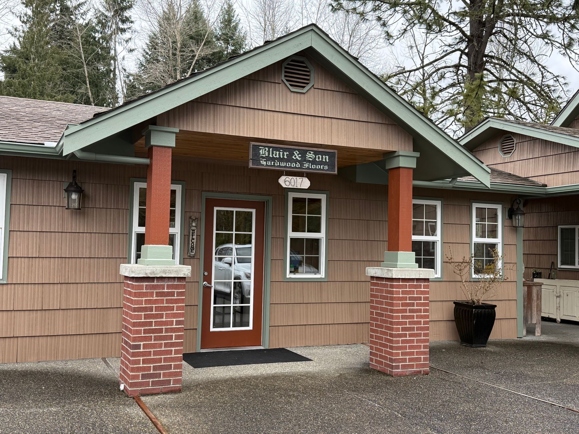 Brown-shingled building entrance with brick-based pillars, a wood-framed glass door, and green-trimmed gabled roof.
