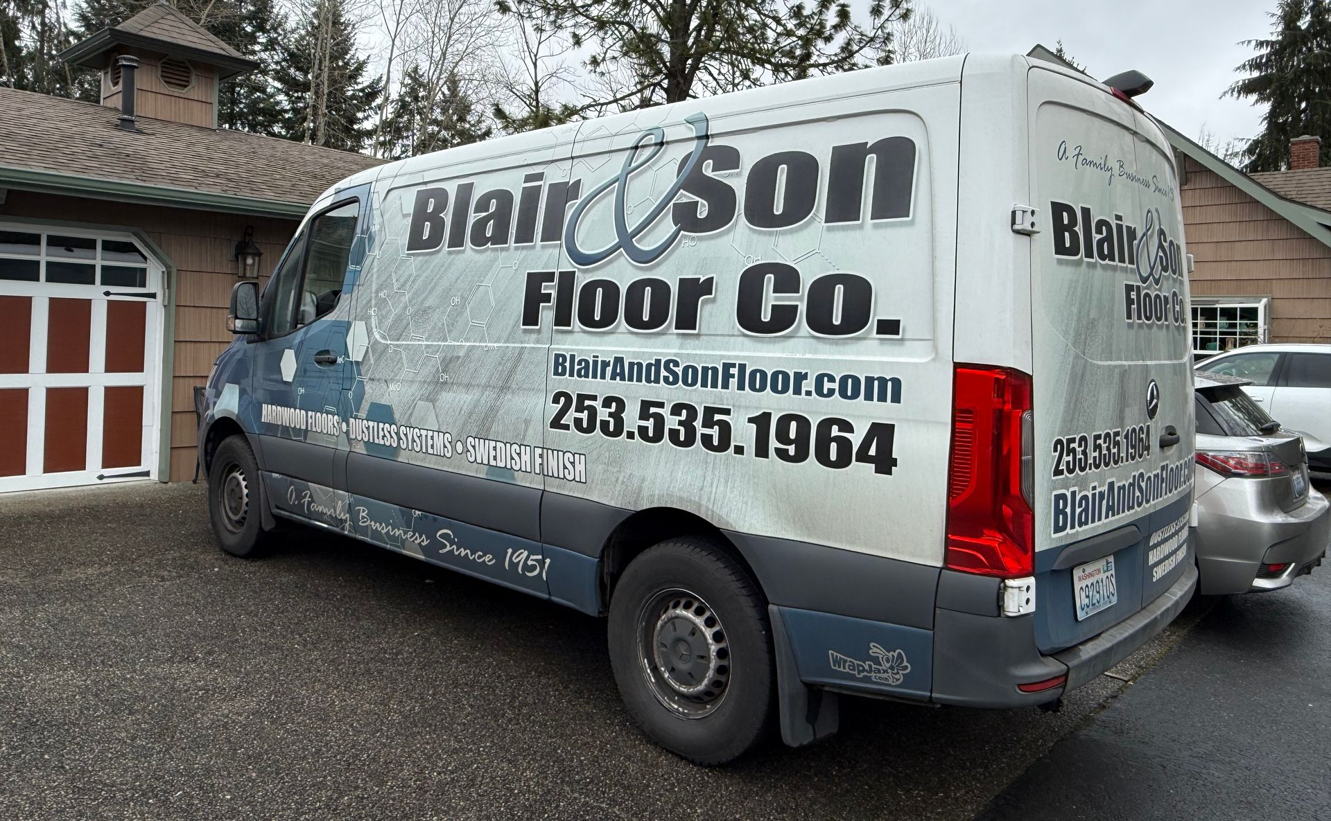 A white work van for Blair & Son Floor Co. parked on a gravel driveway in front of a brick building.