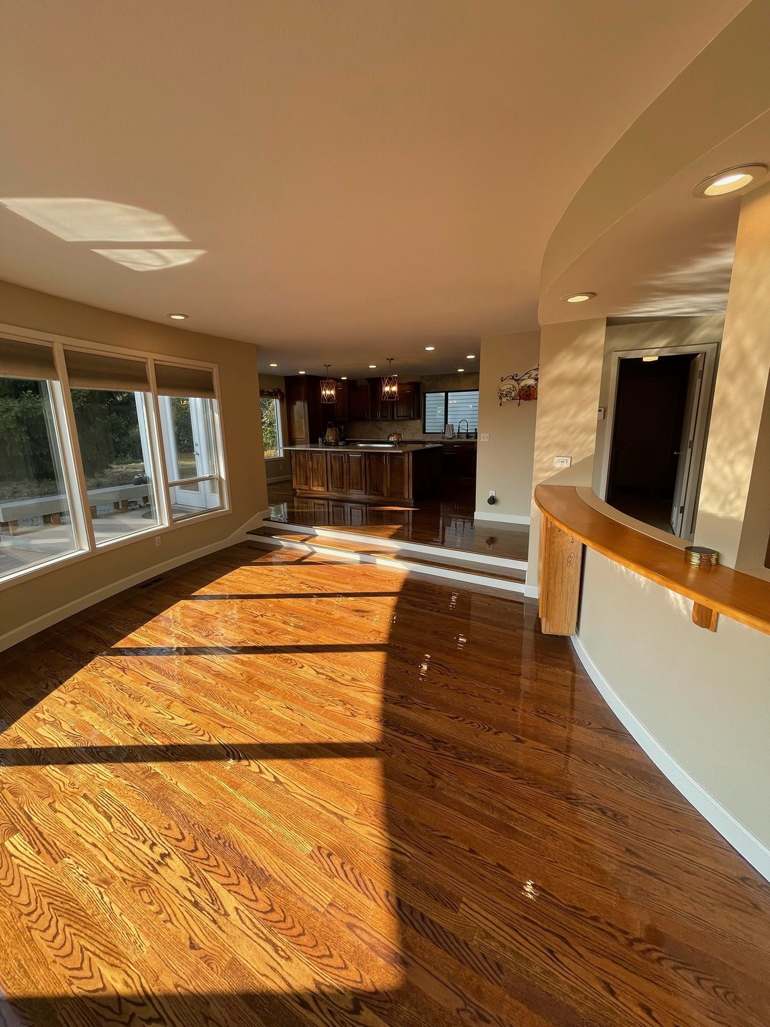 A bright, open-plan living room with polished hardwood floors, large windows, and a kitchen visible in the background.