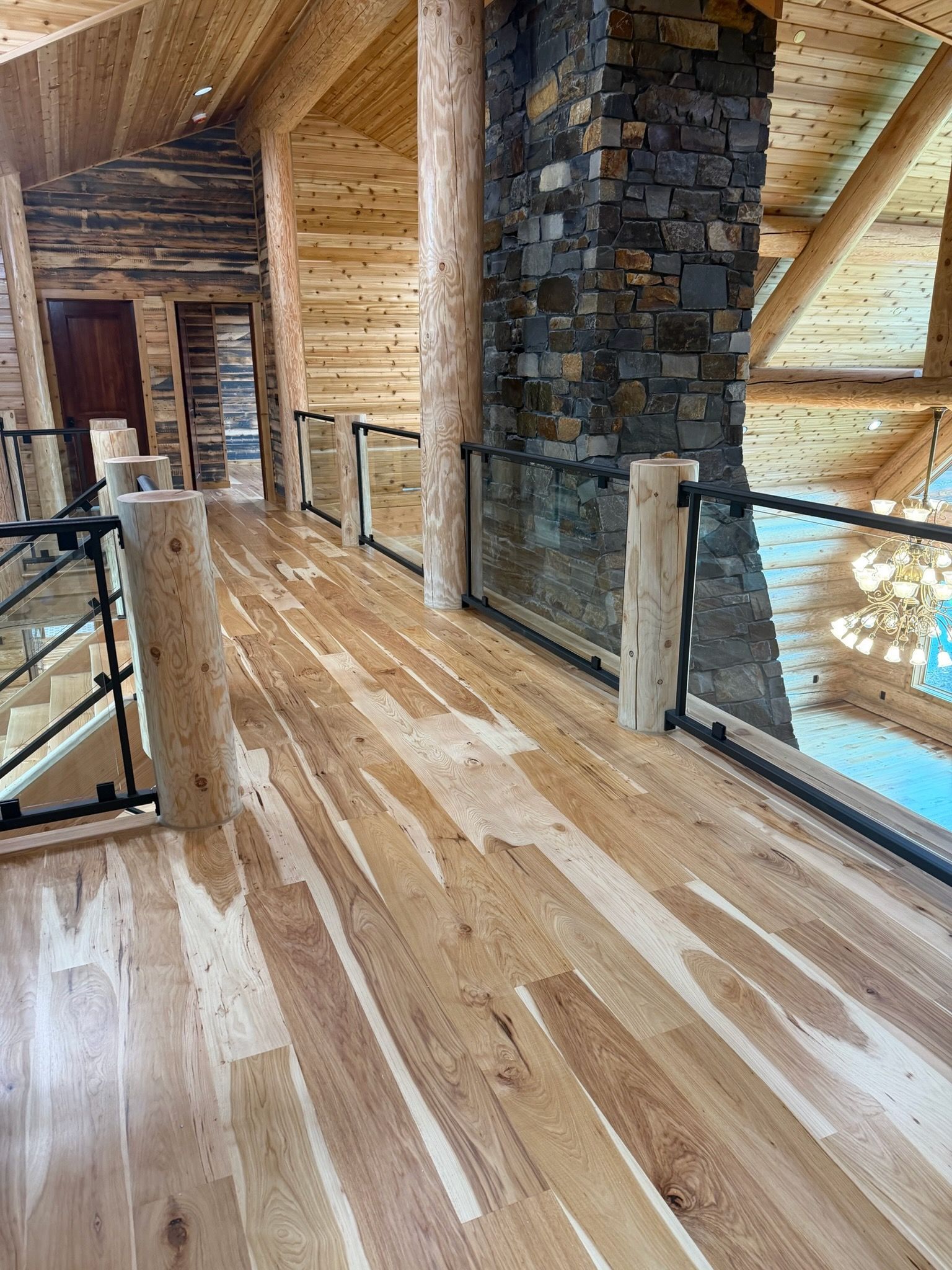 An elevated wood-floored walkway with rustic log posts and glass railings, overlooking a bright, open-plan lodge room.