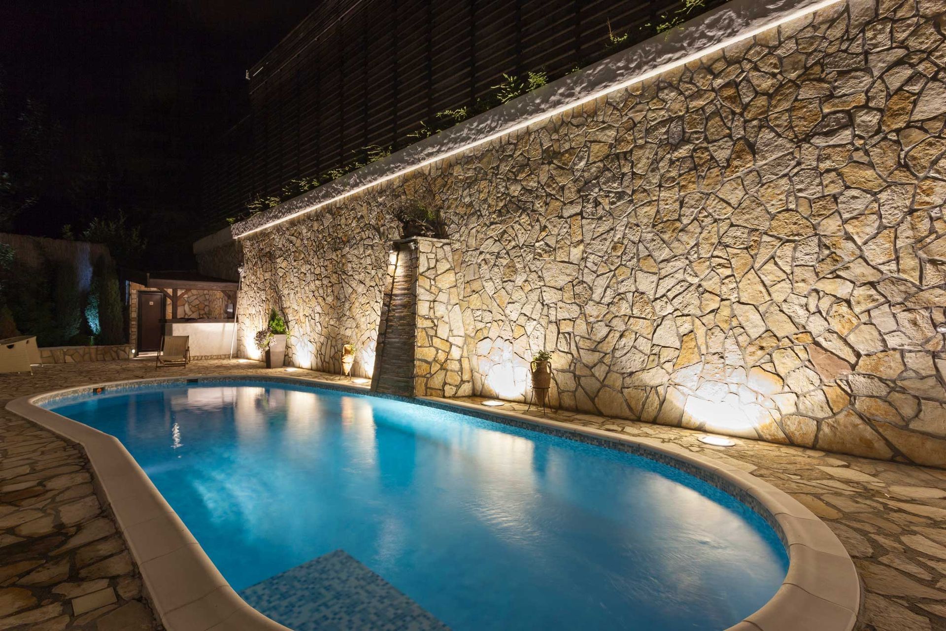 A large swimming pool is lit up at night in front of a stone wall.