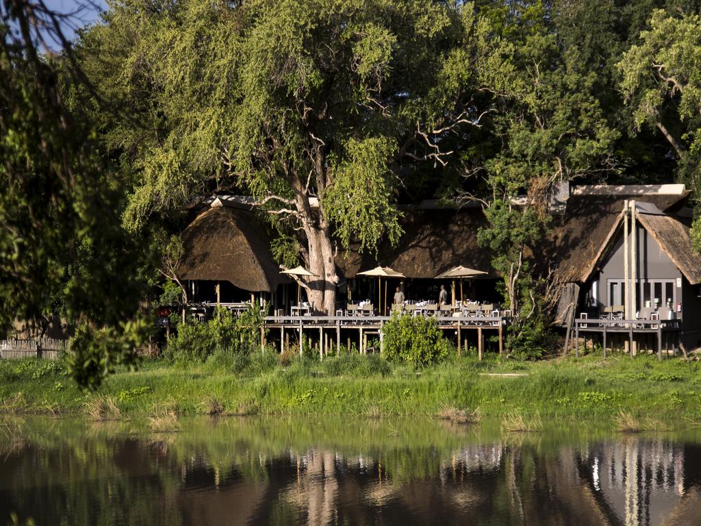 A house with a thatched roof sits next to a body of water surrounded by trees.
