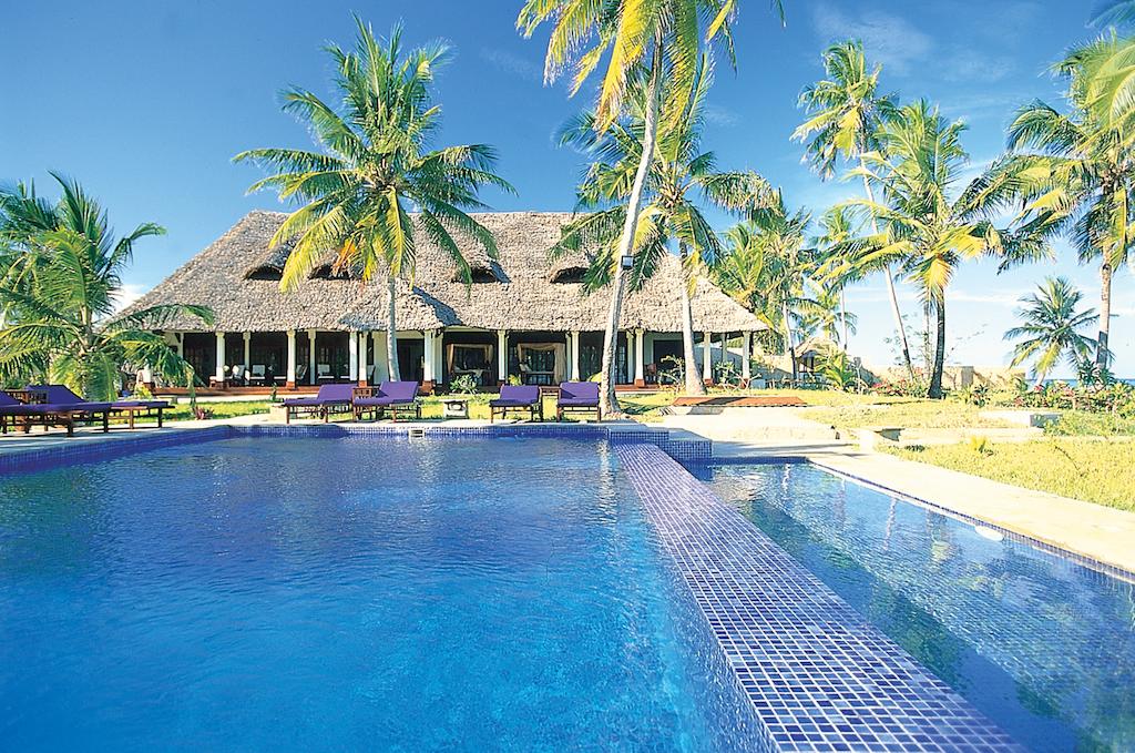 A large swimming pool in front of a thatched house surrounded by palm trees.