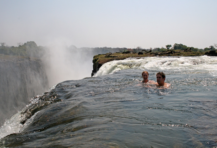 Two people are swimming in the water near a waterfall.