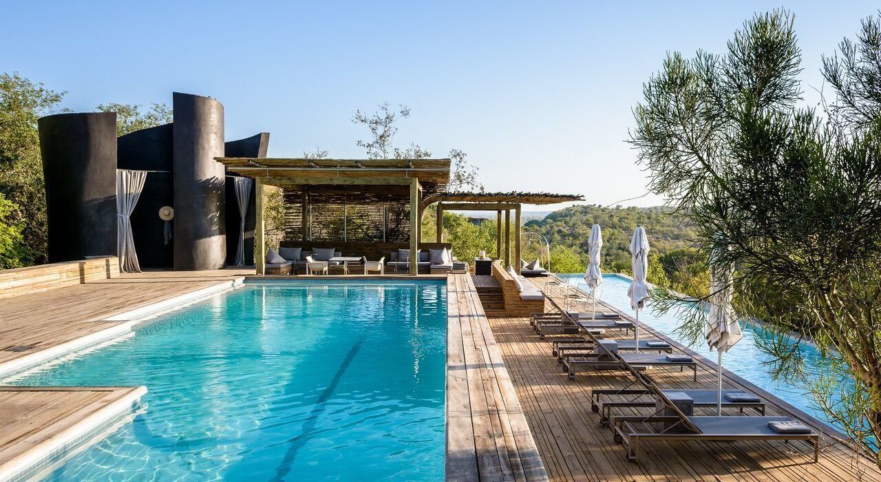 A large swimming pool surrounded by chairs and umbrellas in front of a house.