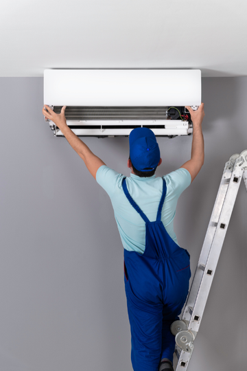 A man is standing on a ladder fixing an air conditioner.