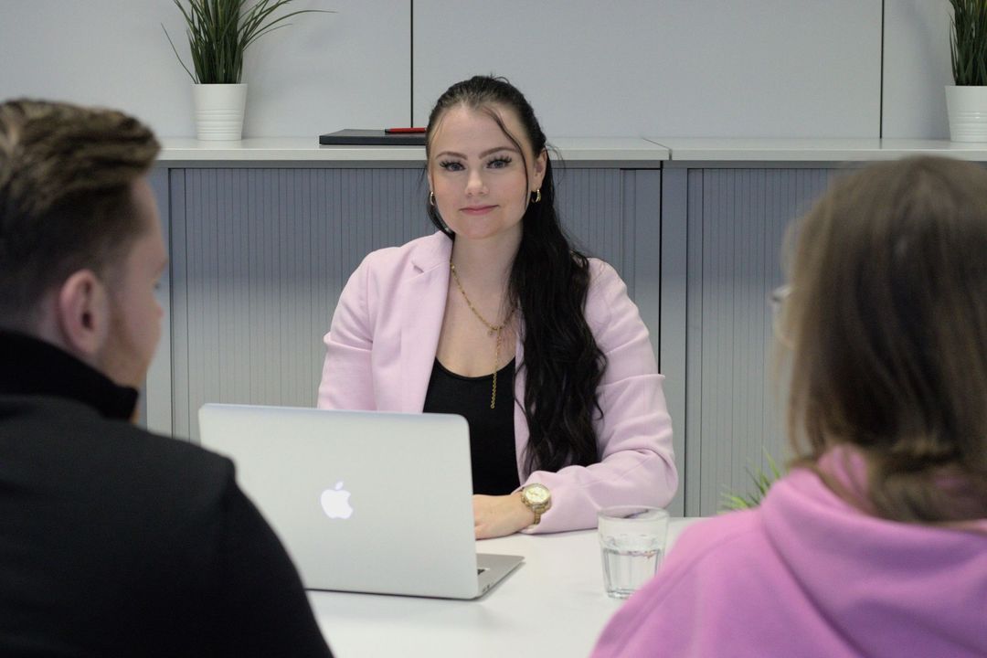 Eine Frau sitzt mit zwei Personen und einem Laptop an einem Tisch.
