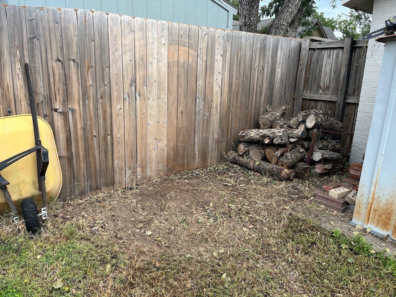 A wooden fence surrounds a pile of logs in a backyard.