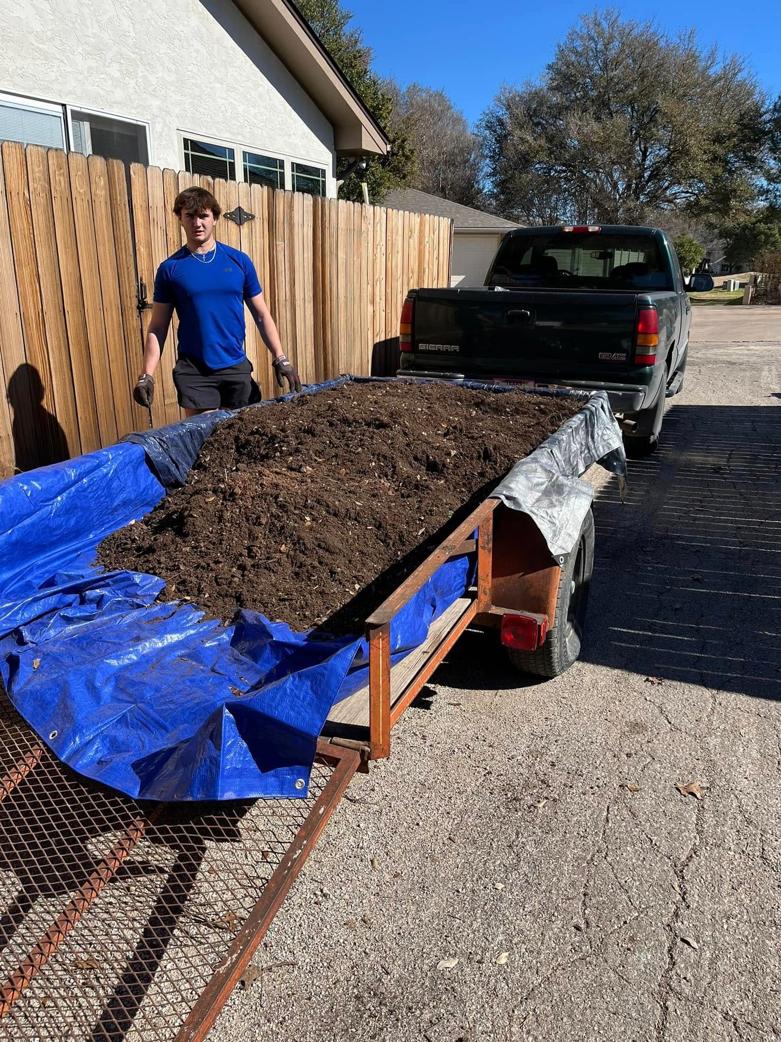 A man is standing next to a truck with a trailer full of dirt.