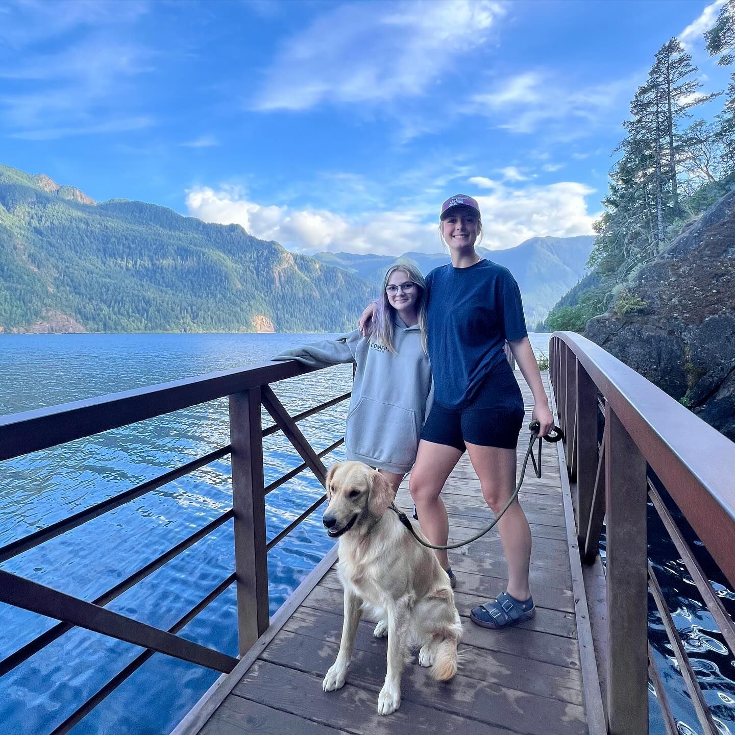 Woman and girl with golden retriever on a bridge overlooking a lake and mountains.