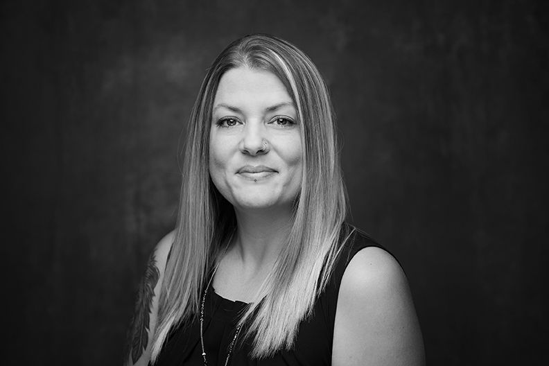 Woman with long blonde hair smiles, wearing a black sleeveless top, against a dark backdrop.
