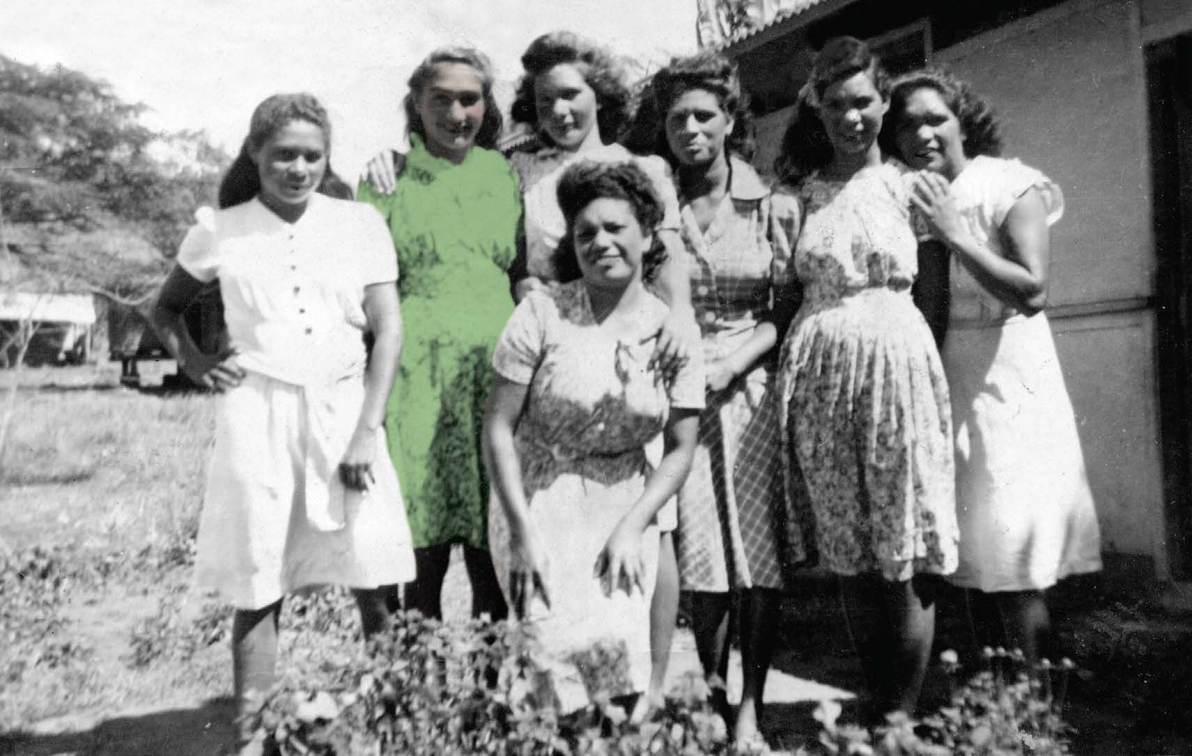 Group of seven women in dresses posing outdoors near a house, some smiling.