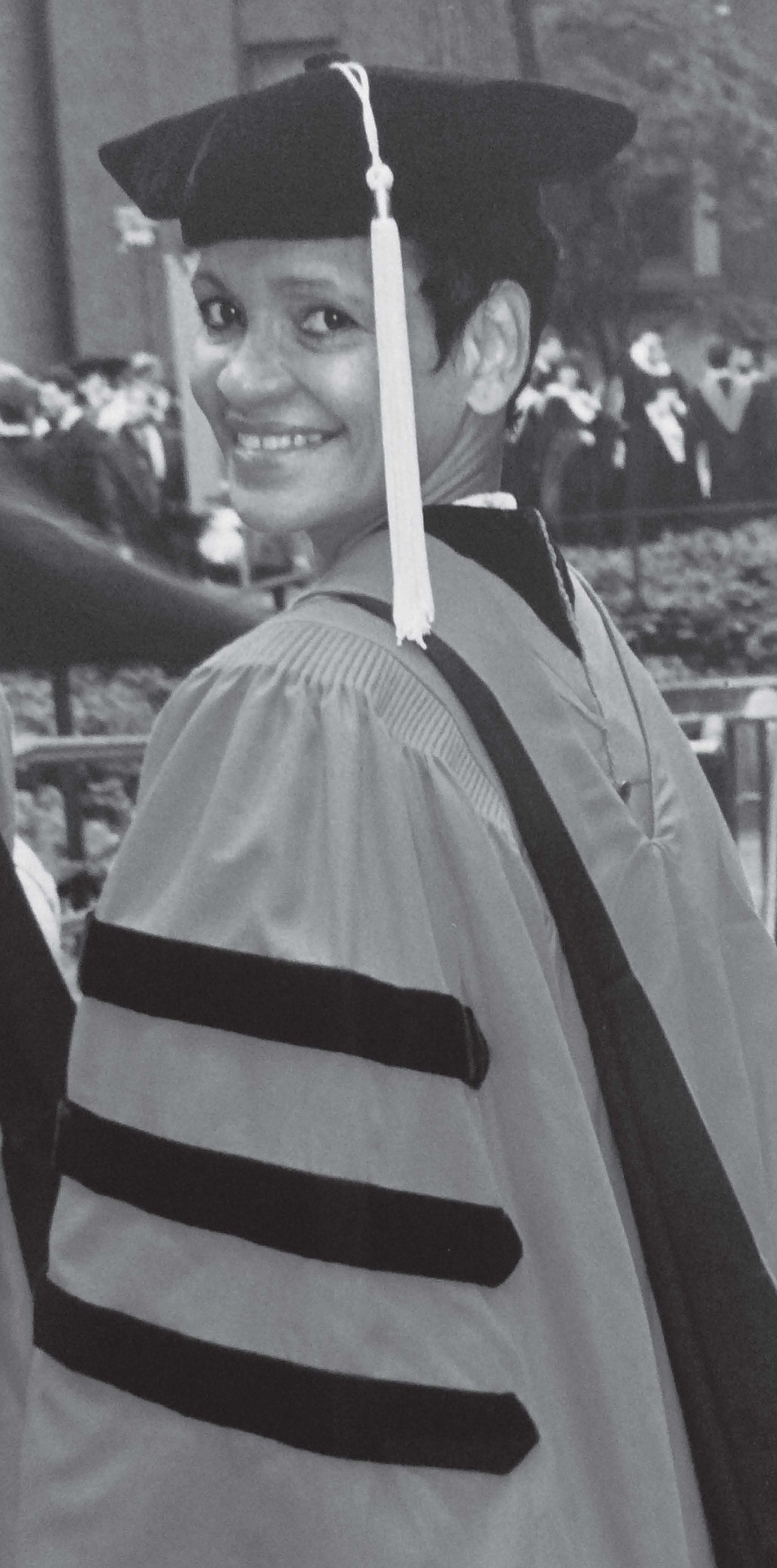 Person in graduation regalia, smiling over their shoulder. Black cap and robes with dark stripes.