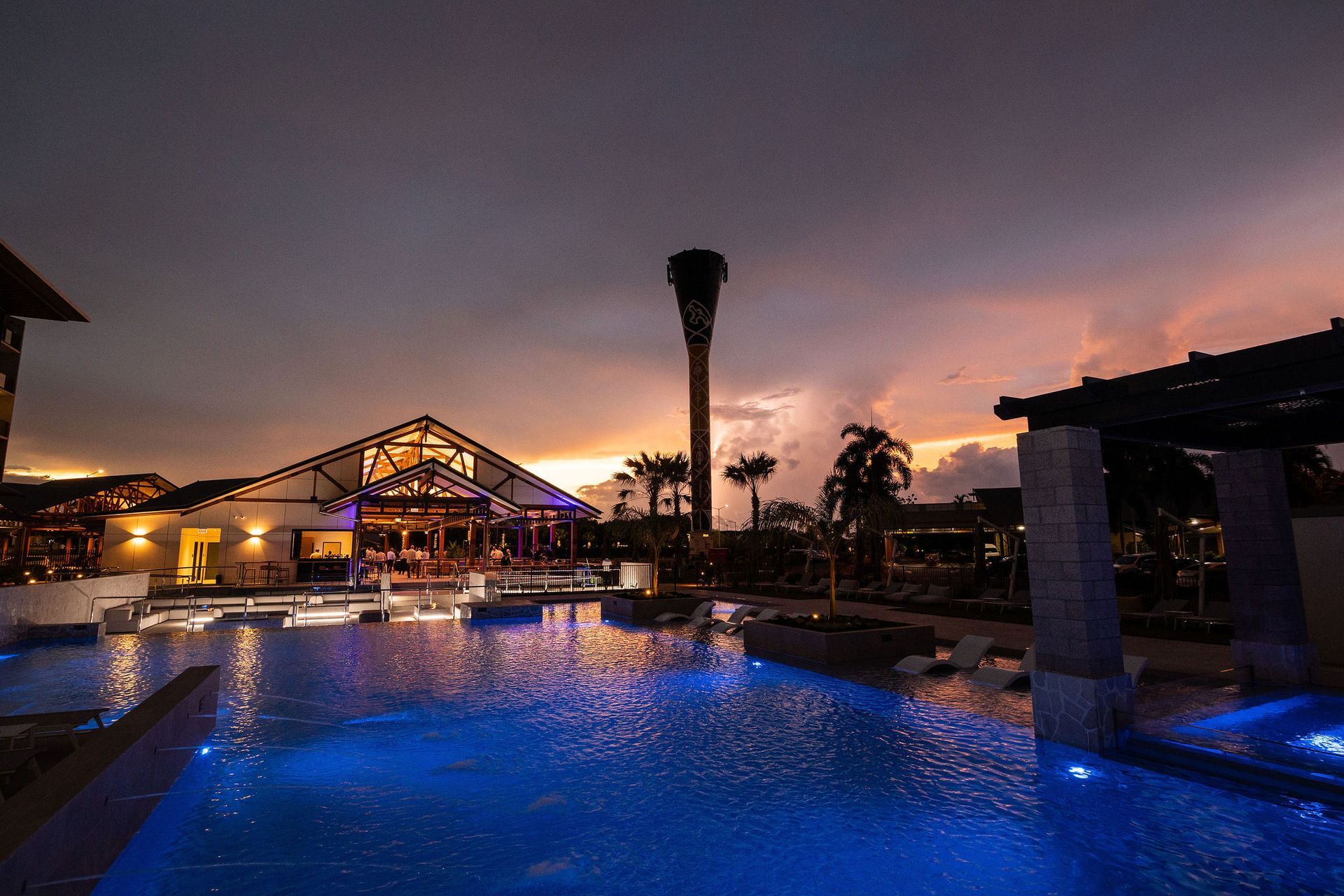 Pool reflecting blue lights, resort building with lights aglow, and palm tree against a colorful sunset.