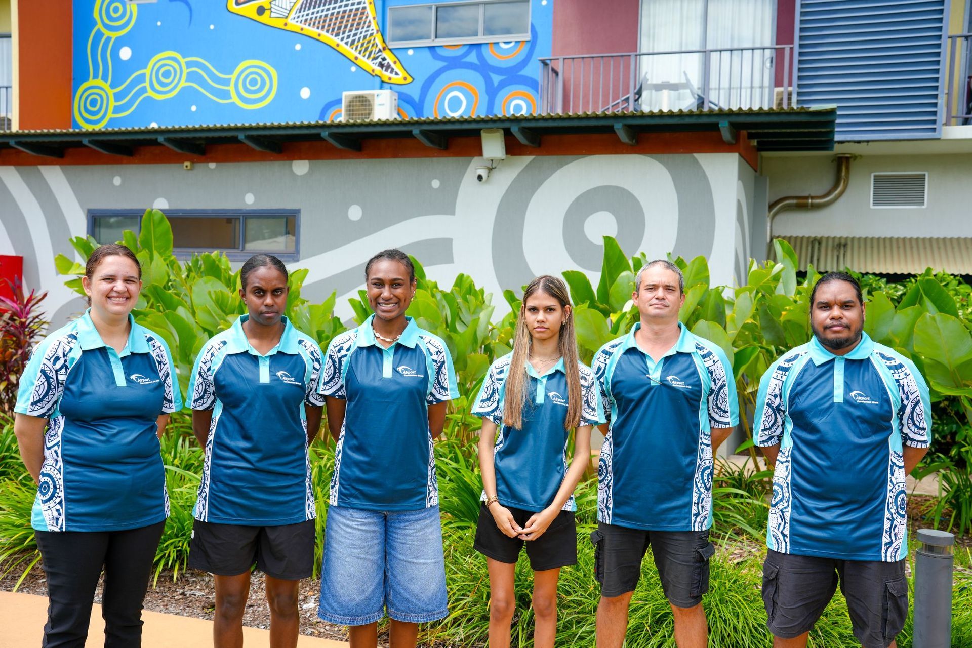 Six people in matching teal and white patterned uniforms stand together in front of a colorful, painted mural.