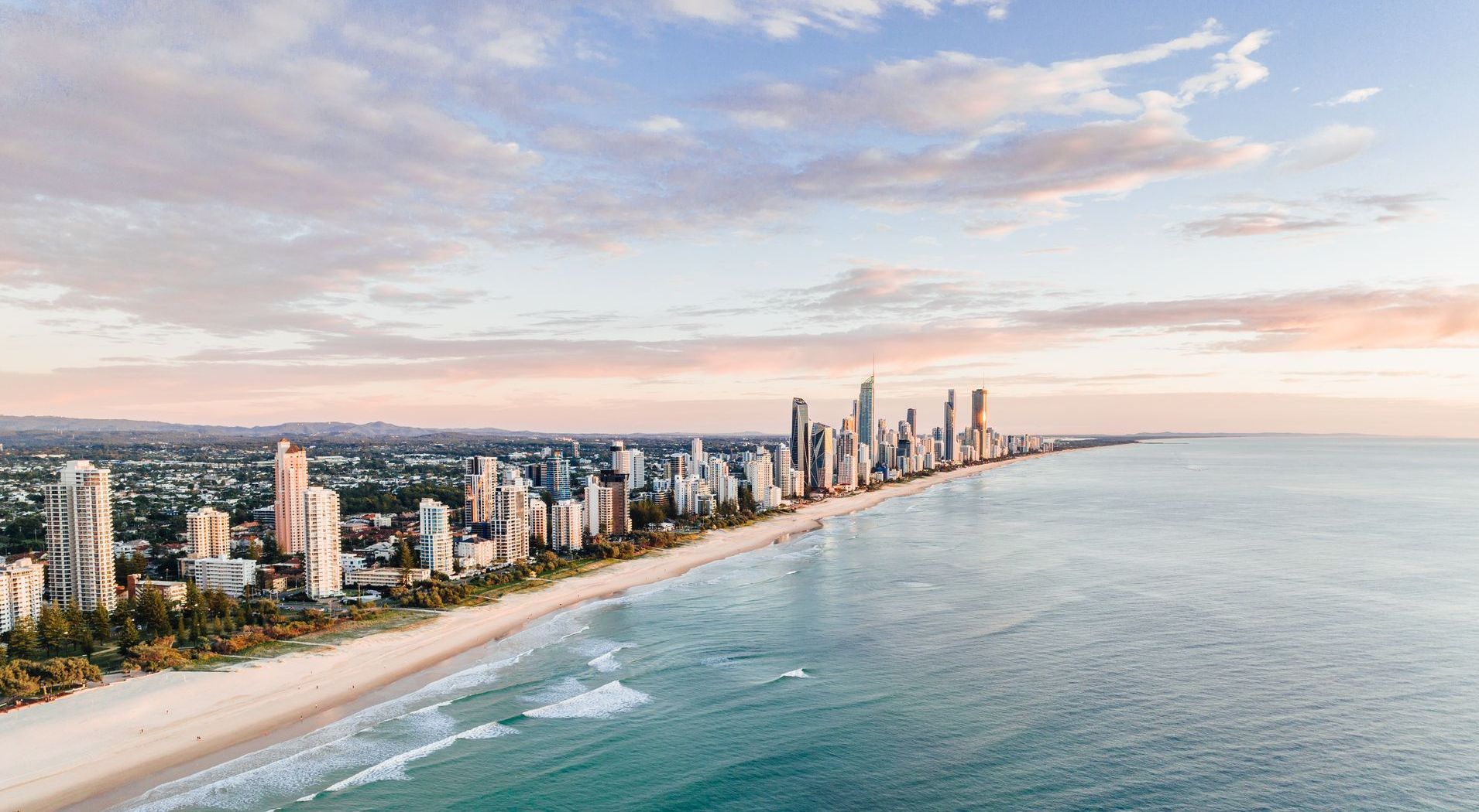 Coastal cityscape with high-rise buildings, sandy beach, and turquoise water under a pastel sky.