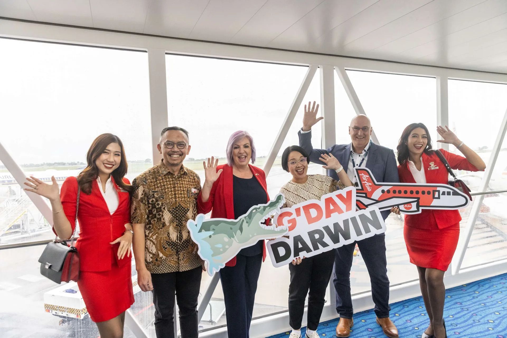 Group of people waving, holding a sign that says "G'Day Darwin" in a brightly lit airport hallway.