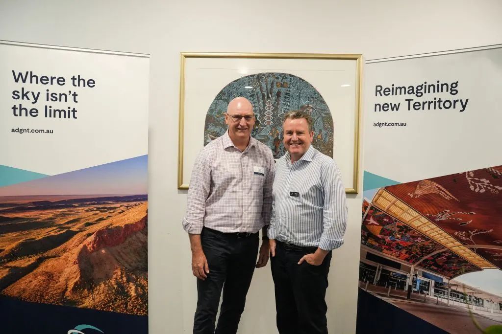 Two men pose in front of artwork and banners in an office. One smiles, the other looks at the camera.