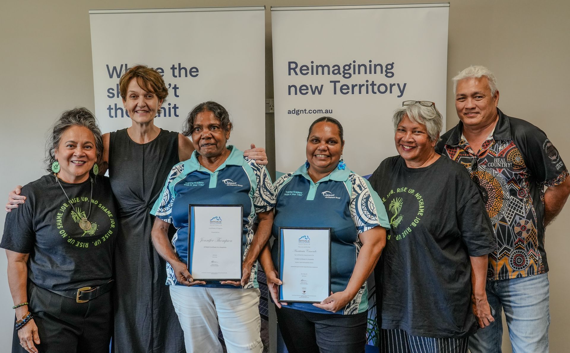 Group of people, including two women holding framed certificates, posing in front of banners; indoors.