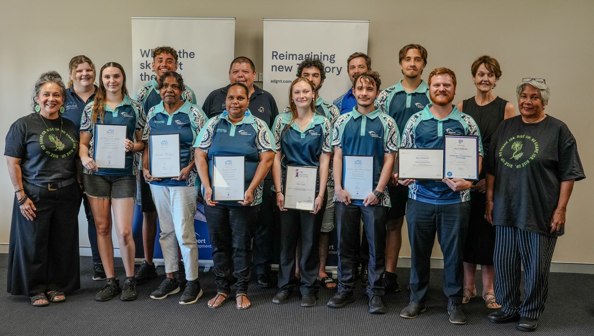 Group of people holding certificates; some smiling. Indoor setting with banners in background.