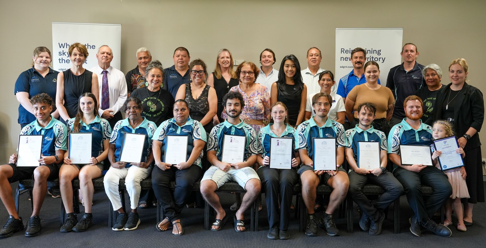 Group photo of award recipients with certificates and supporting staff. Indoors, smiles, and various skin tones.
