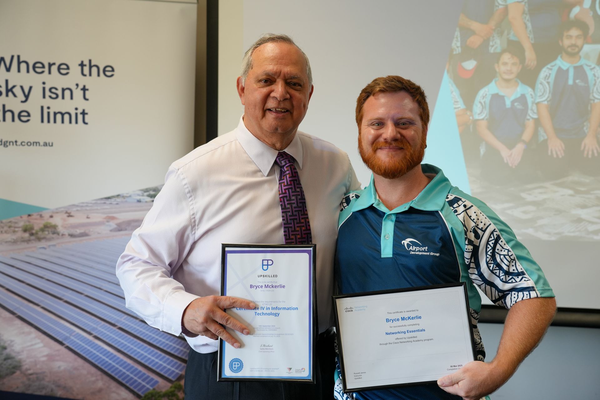 Two men holding certificates, smiling. One in a suit, the other in a polo shirt, standing in an office.