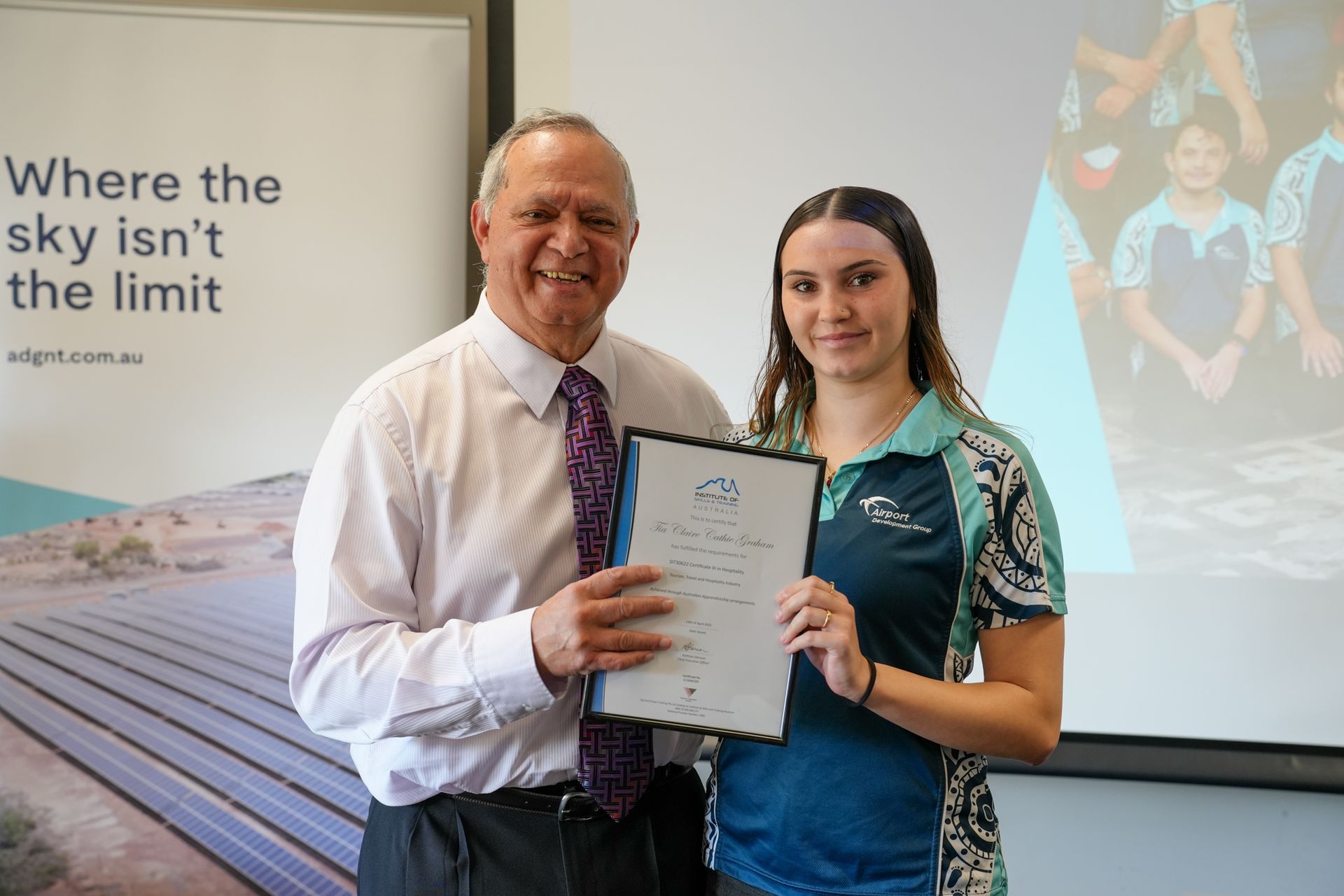 Man presenting a framed document to a smiling young woman. They stand in a room with a presentation screen.