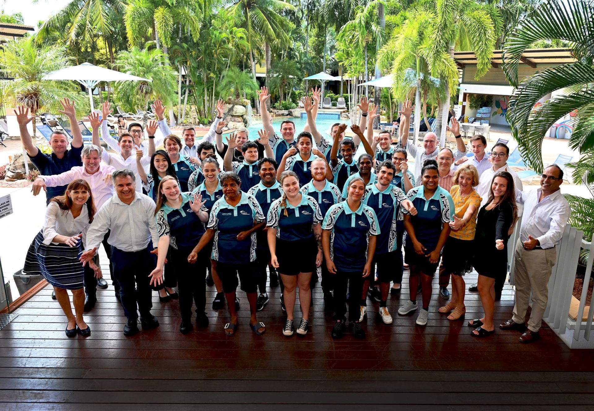Group of people smiling with arms raised on a wooden deck outdoors, possibly at a resort.