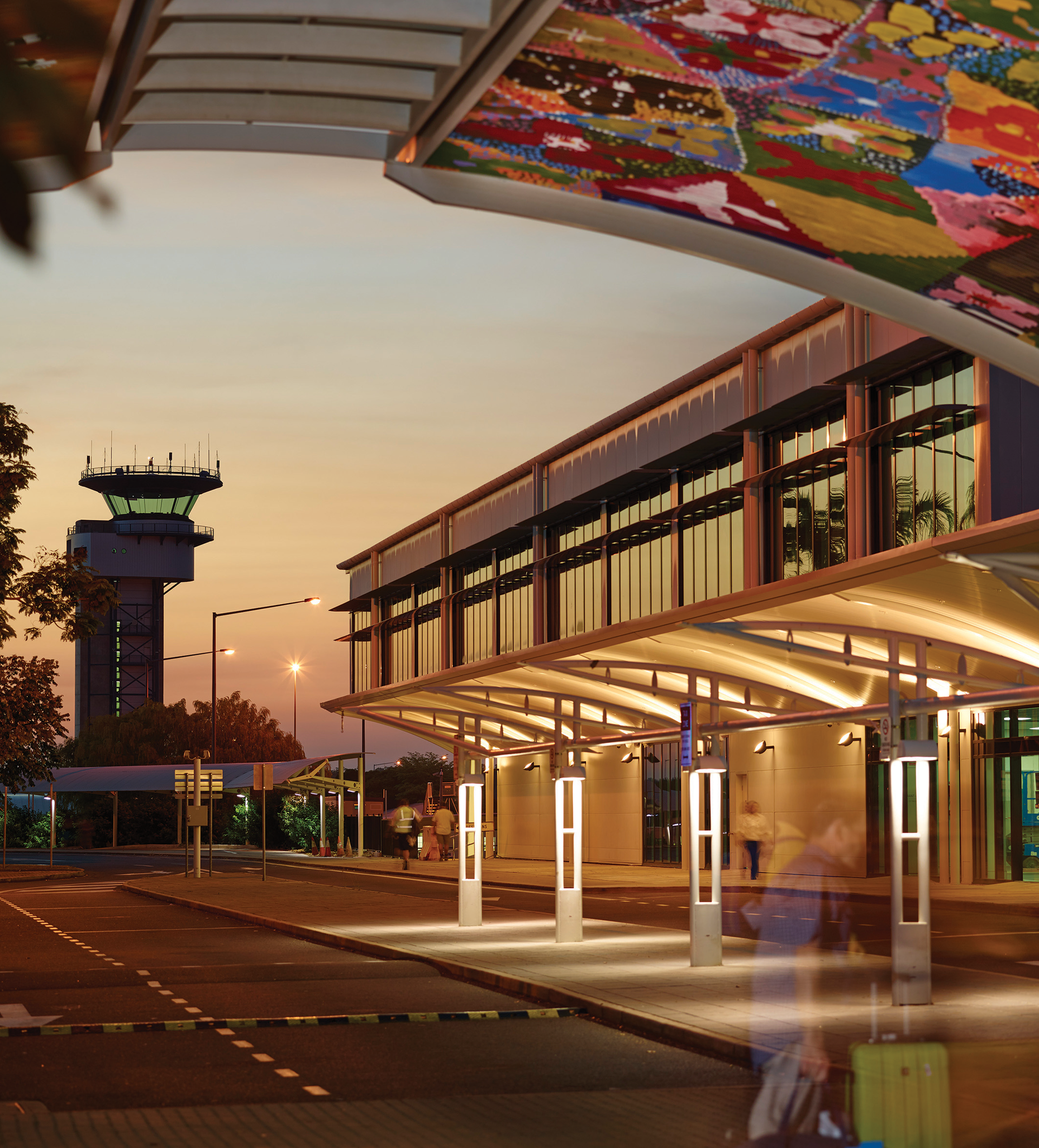 Airport exterior at dusk with control tower, covered walkway, and blurred figures.