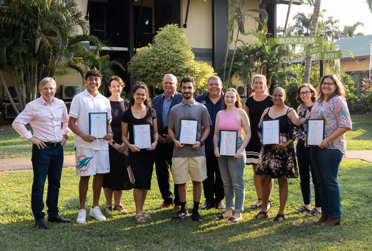 Group of people holding certificates, smiling outdoors on a lawn. Trees and building in the background.