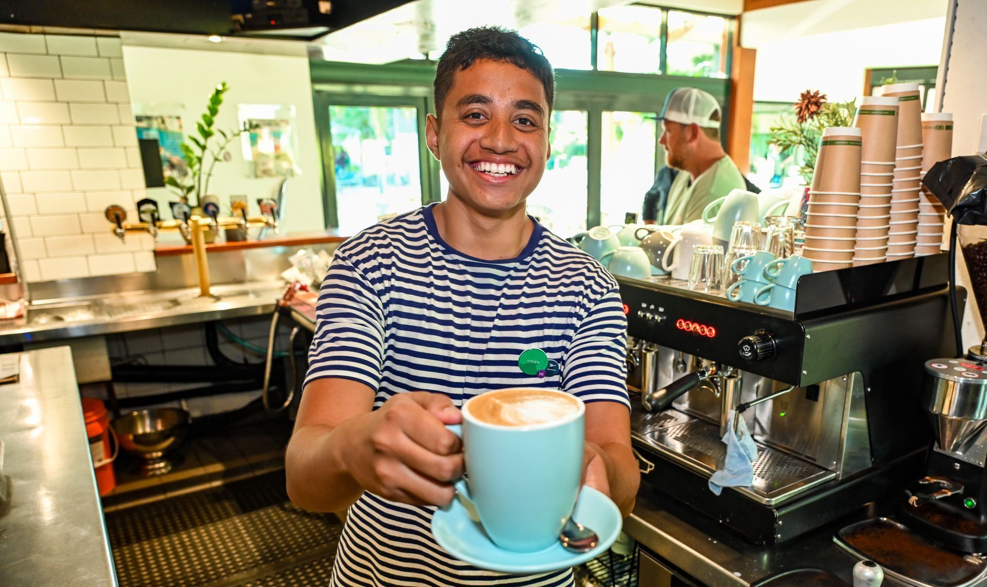 Smiling barista offers a latte in a blue mug; cafe setting.