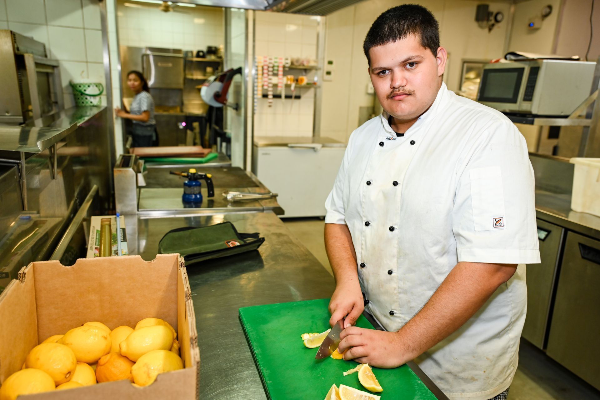 Chef cutting lemons in a commercial kitchen; a box of lemons nearby, another person in the background.