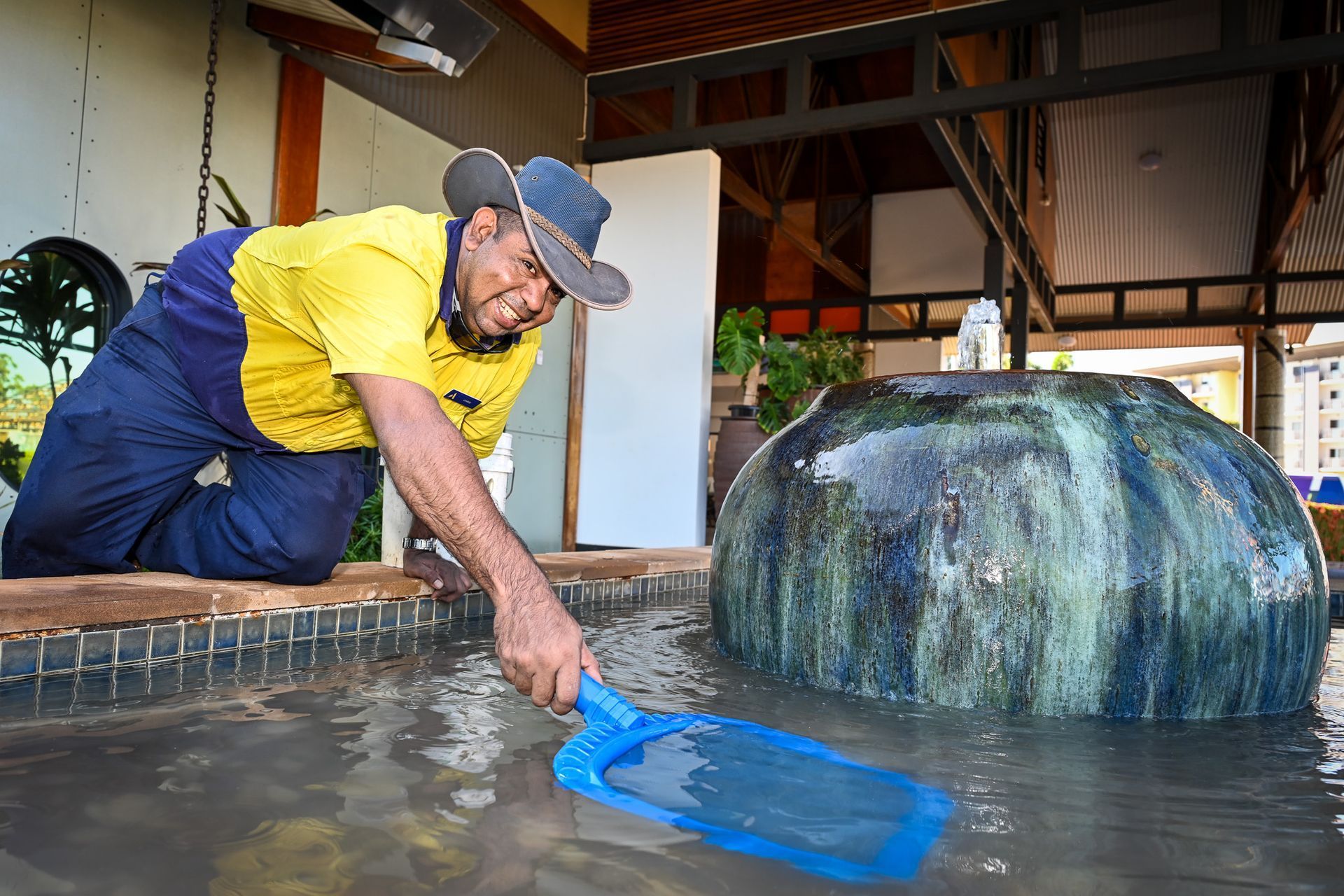 Man cleaning a fountain with a net; outdoors, wearing work clothes and a cowboy hat.