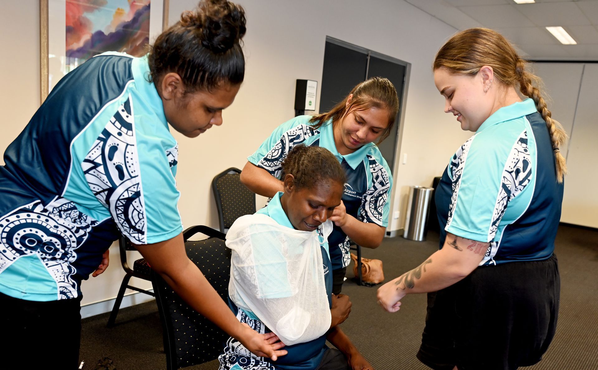 Four people in teal shirts practice first aid on a person with an arm injury, indoors.