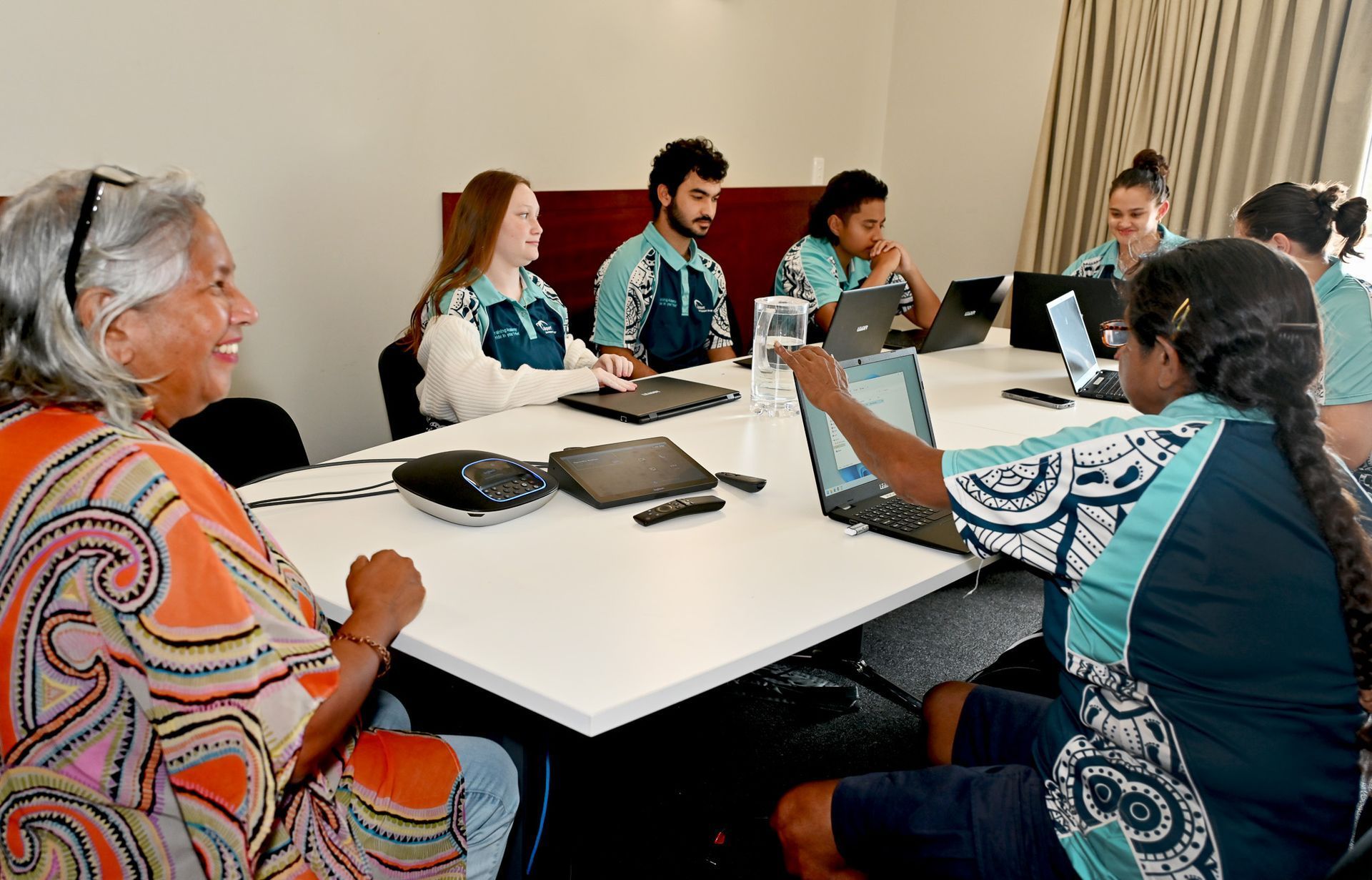 Group of diverse people in a meeting, some using laptops, smiling. Bright room, white table, casual attire.