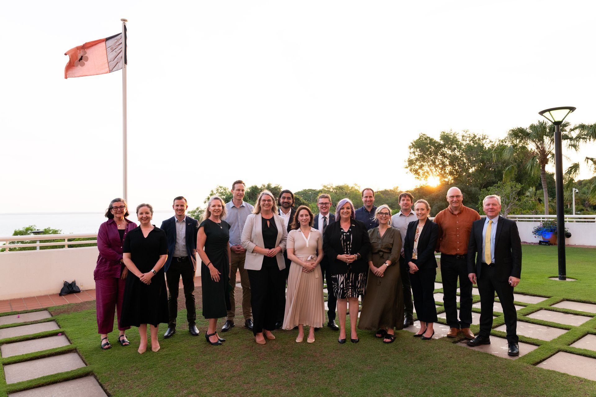 Group of people posing outdoors with a flag.  Sun setting in the background.