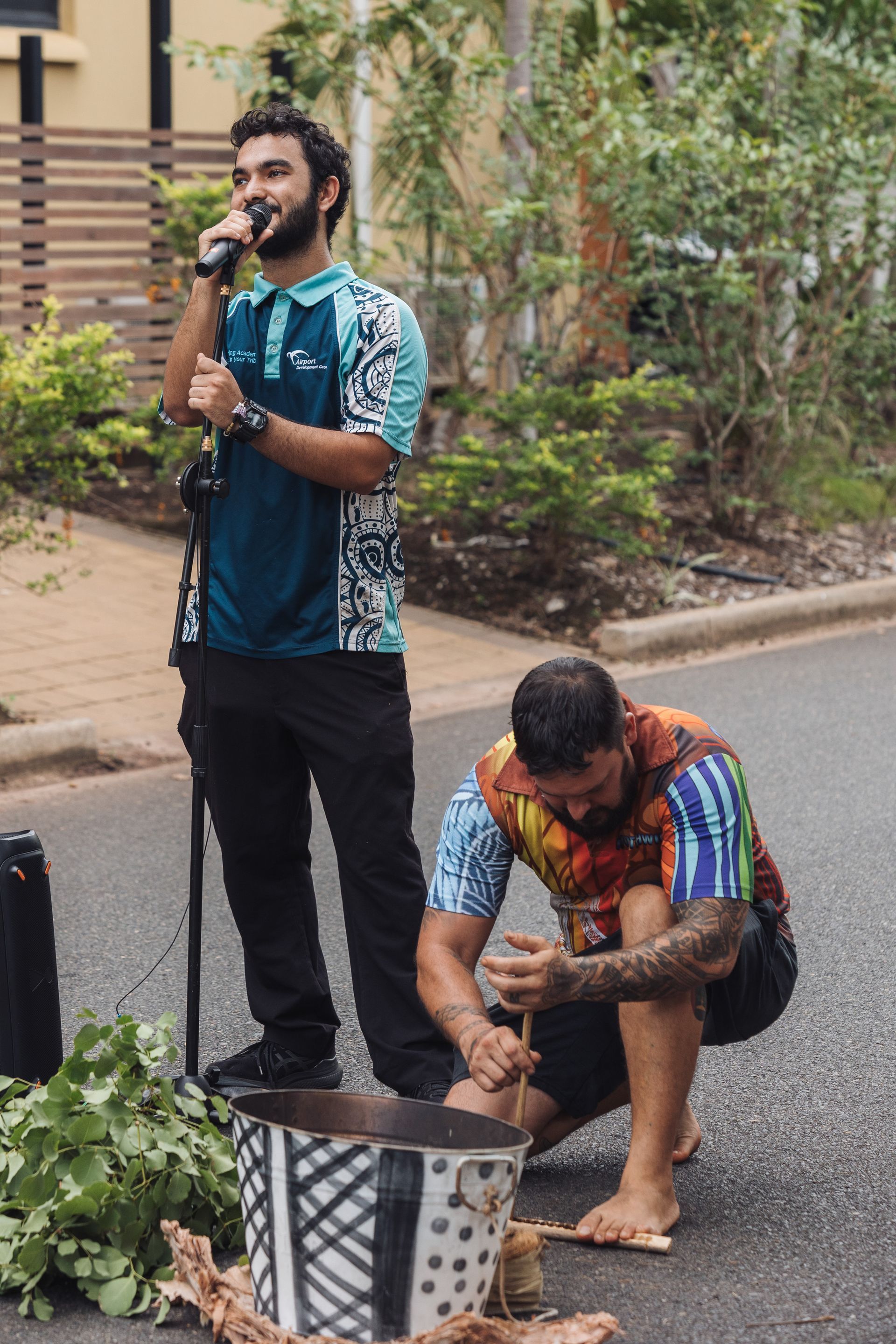 Two men, one speaking into a mic, the other stirring a pot. Outdoors, street setting.