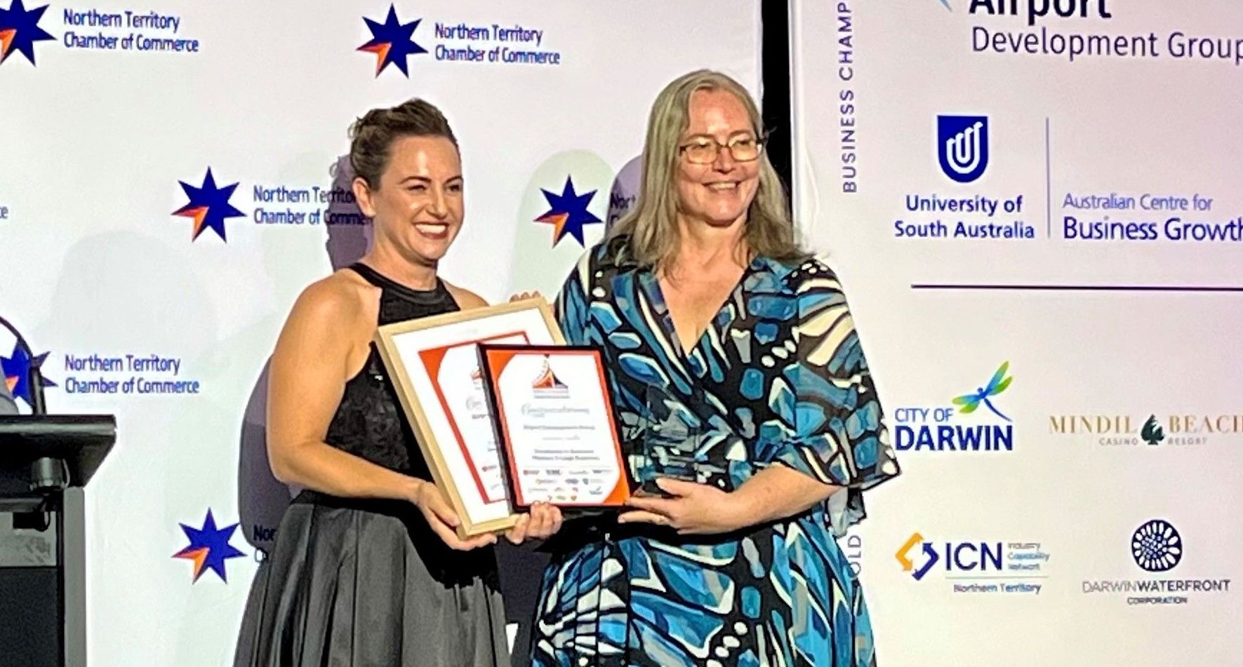 Two women smiling, holding awards on stage with logos in background.
