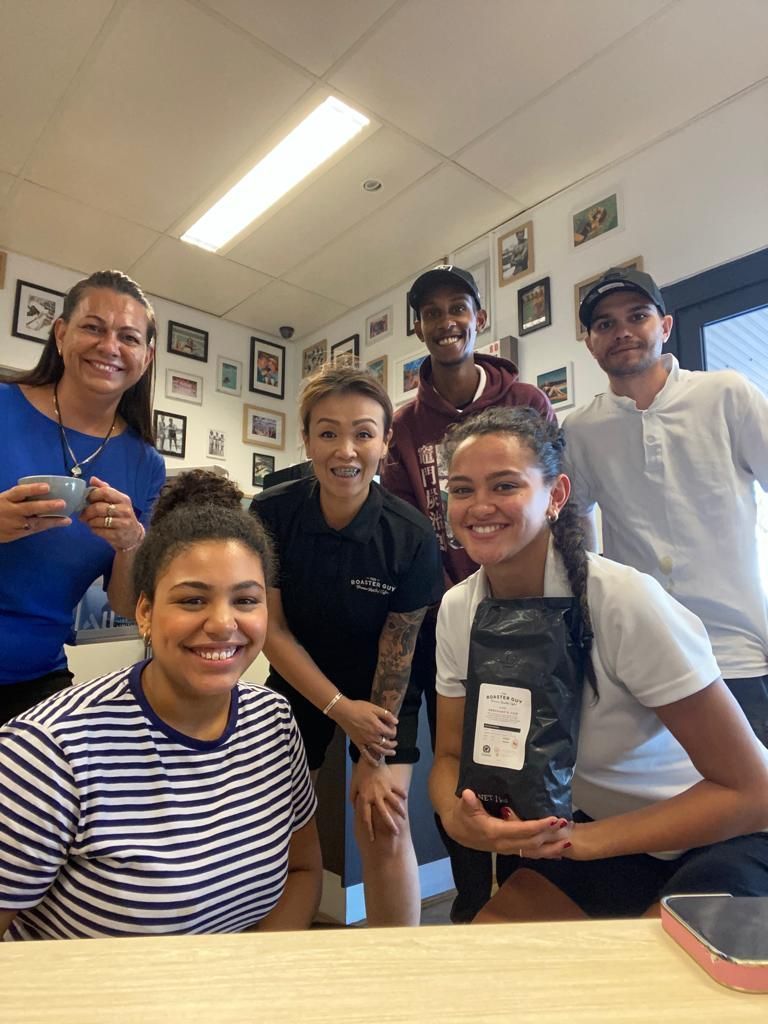 Group of seven people in a coffee shop, smiling. One holds a bag of coffee beans.