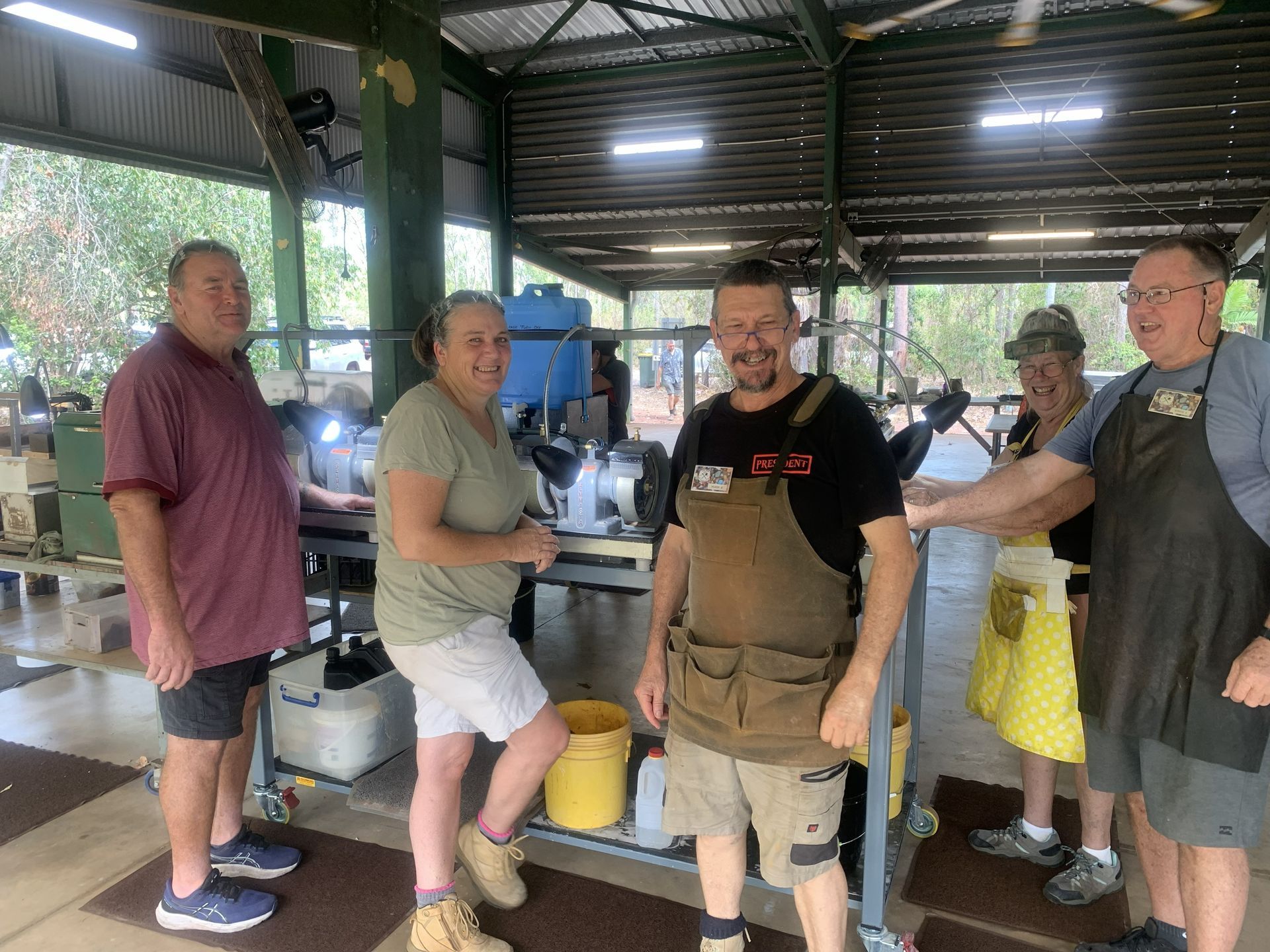 Four people standing in a covered workshop area, smiling and posing near grinding equipment and tools.