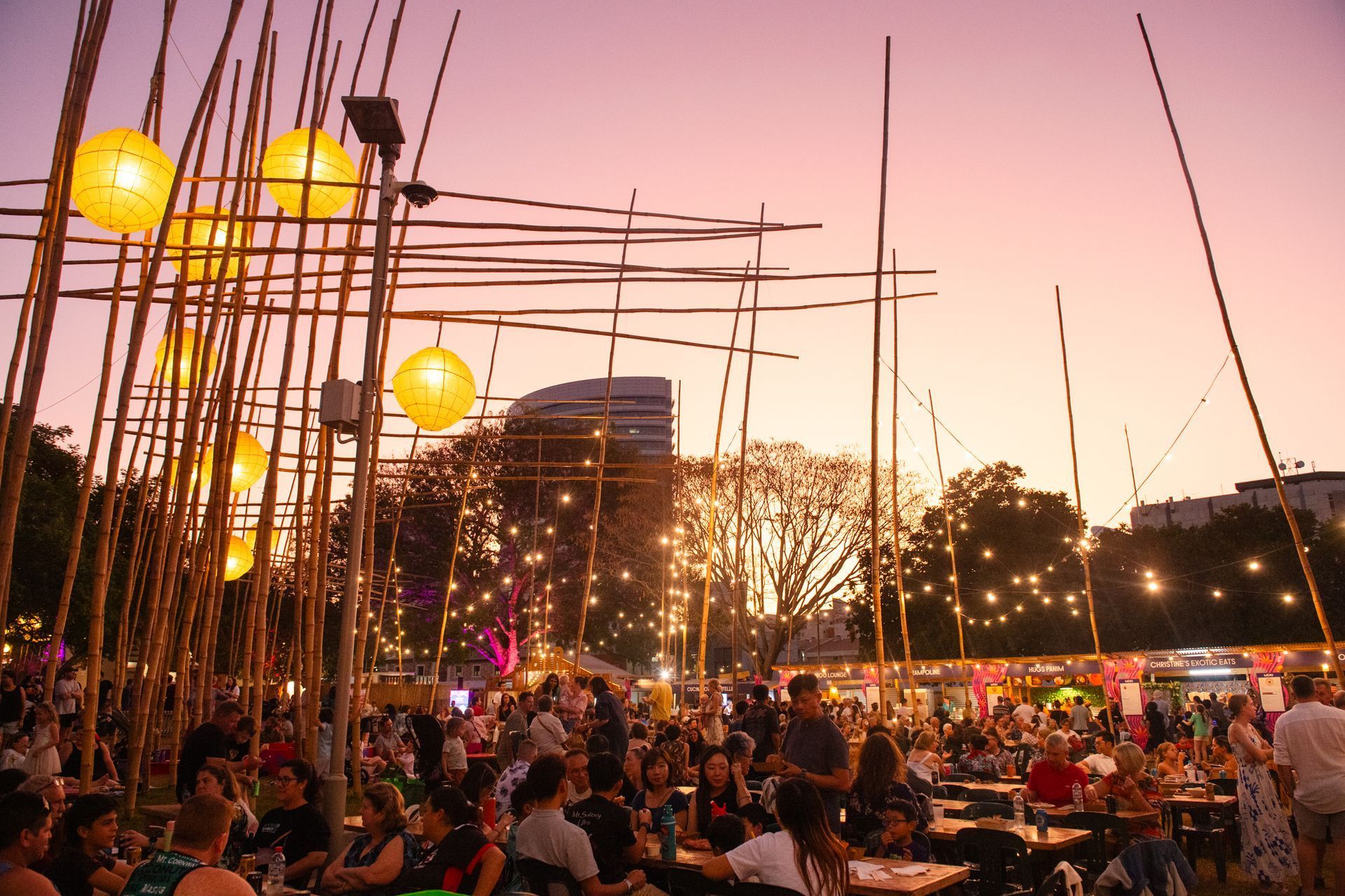 Crowd at outdoor festival under bamboo structures with lanterns at sunset.